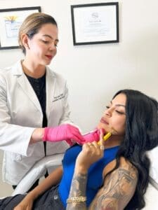 Specialist Wearing A White Coat Black Uniform And Pink Gloves Applies Radiesse Dermal Fillers Injection To A Woman Holding A Facial Roller To Her Face