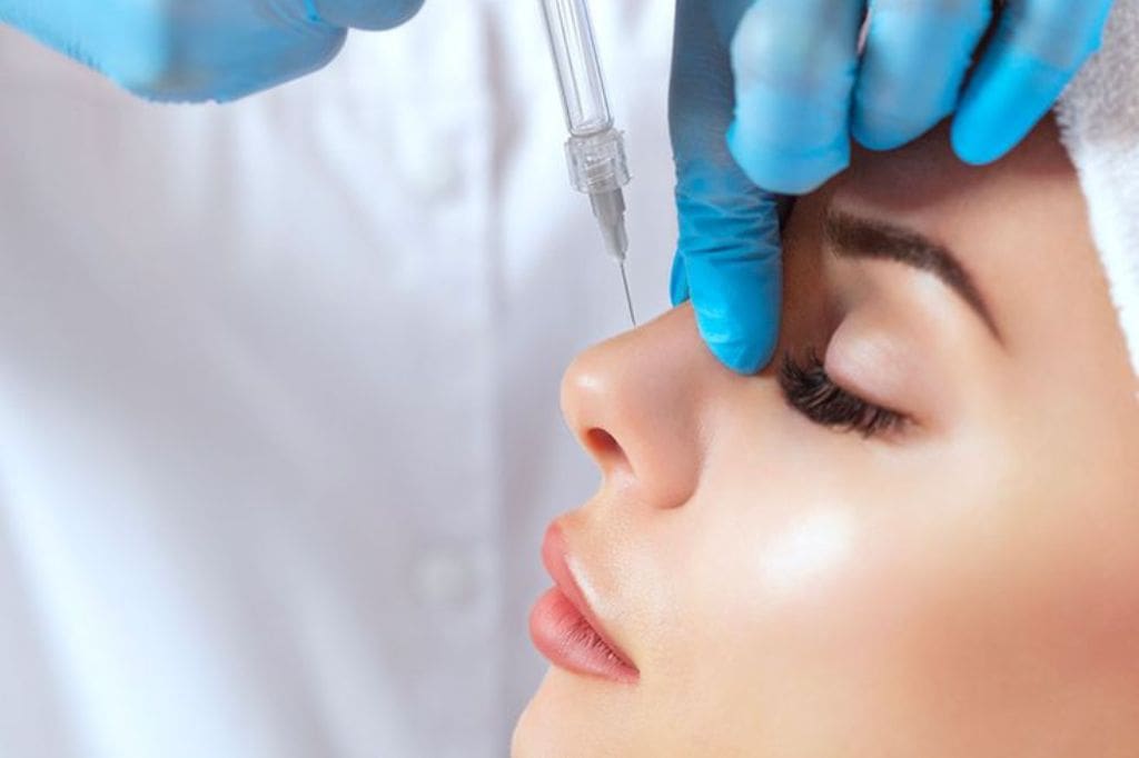 Patient Wearing Spa Headband Receiving Botox Dermal Filler On Her Nose With A Syringe By A Specialist Wearing A Doctor Coat And Blue Gloves