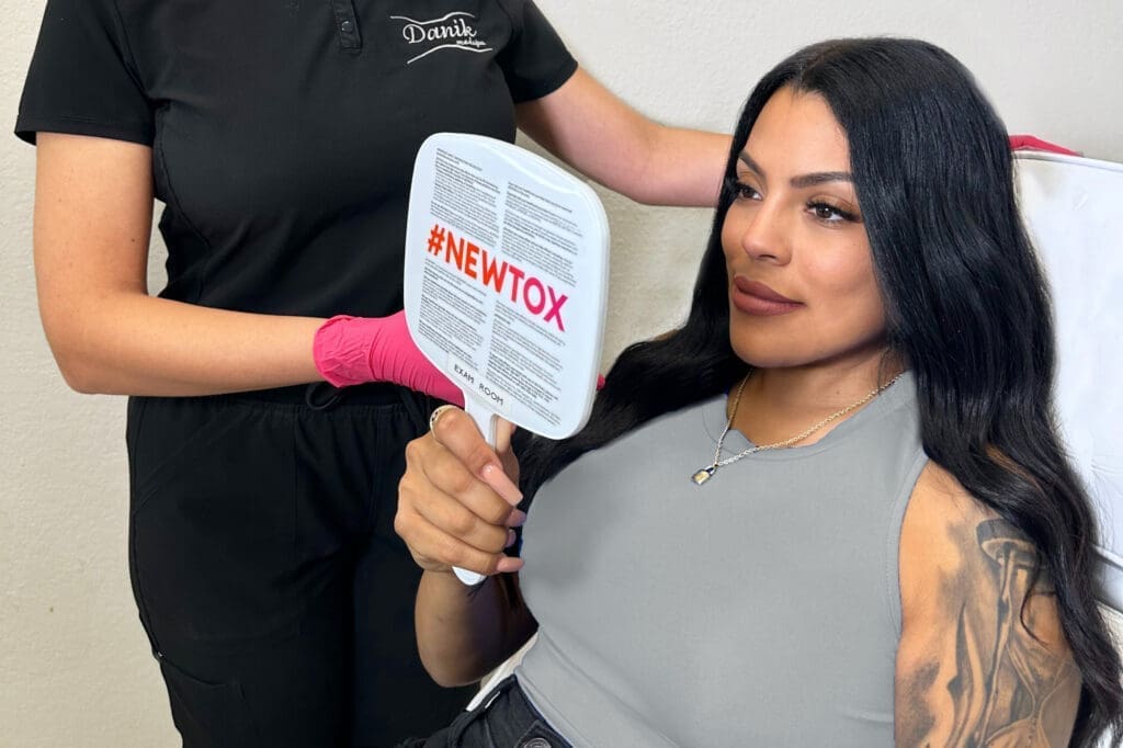 Woman In Gray Shirt Sees Results Of Jeuveau Treatment In A Handheld Mirror With Specialist Behind