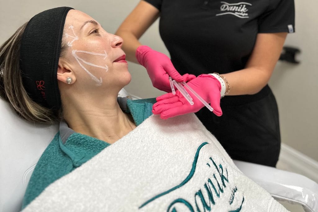 Specialist Wearing Black Uniform And Pink Gloves Show PDO Threads To Woman Patient With White Markings On Her Face To Outline The Treatment